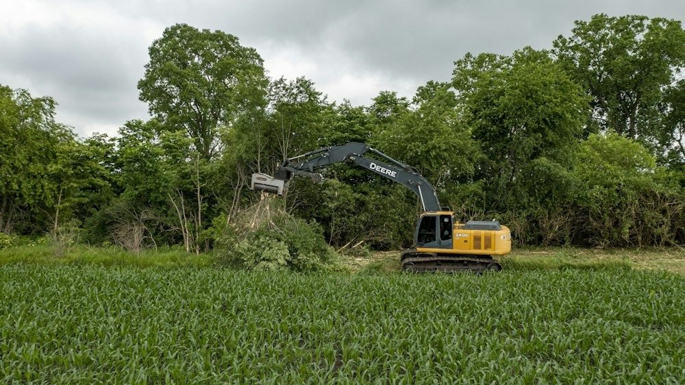 A mini excavator pulling stumps and brush on a partly cleared wooded lot