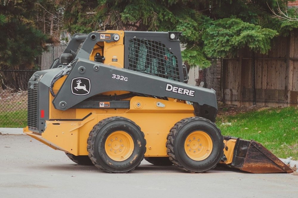A compact track loader parked at the edge of a field beside a pine tree line